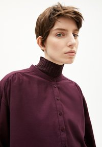 Camicetta bordeaux con colletto alto a coste. Presenta una chiusura con bottoni, una vestibilità comoda e un materiale in cotone liscio. Design elegante e minimalista.