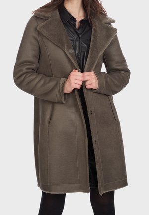 Vrouw draagt een knielange olijfgroene schapenvachten jas met brede revers over een zwarte blouse met knopen, houdt de jas bij de borst dicht.