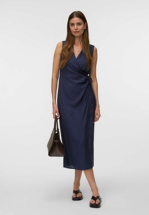 Mujer con cabello largo y castaño que lleva un vestido cruzado sin mangas en azul marino, llevando un bolso marrón y sandalias negras con tacón.