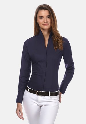 Camicia button-up a maniche lunghe di colore blu navy con colletto alto, vestibilità slim e trama liscia, abbinata a pantaloni bianchi e una cintura nera.