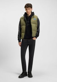 Chaleco acolchado verde oliva sobre una sudadera con capucha negra, combinado con jeans negros ajustados y zapatillas negras, con un logotipo en el chaleco.