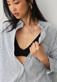 Camisa de botones a rayas en azul claro y blanco, llevada abierta sobre un sujetador negro. La modelo lleva pendientes grandes de aro de plata. Textura suave.