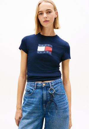 Junge Frau mit glatten, blonden Haaren trägt ein marineblaues Tommy Jeans T-Shirt und hoch taillierte blaue Jeans mit einem Schlüsselanhänger an der Gürtelschlaufe.