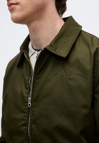 Homme portant une veste zippée vert olive avec le logo "Schott NYC" sur la poitrine et une chemise beige en dessous, tourné vers la gauche sur un fond blanc.