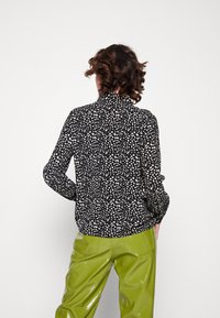 Black blouse with a white polka dot pattern, long sleeves, high collar, and textured fabric, paired with shiny green trousers.