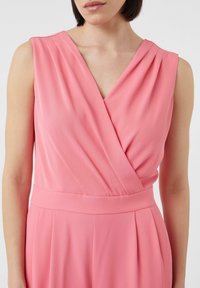 Roze mouwloze jumpsuit met een V-hals, voorzien van geplooide details en een aangesloten tailleband, gemaakt van een gladde stof.