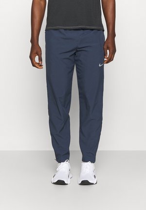 Mann trägt dunkelblaue Nike Jogginghose und weiße Sneakers, steht mit entspannt an den Seiten herunterhängenden Armen vor einem schlichten hellen Hintergrund.