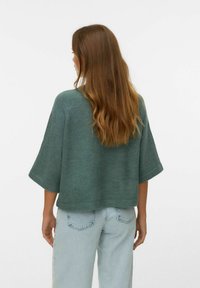 Groene, geknipte gebreide trui met brede mouwen; zachte textuur. Draag met lichtblauwe, high-waisted jeans, bekeken vanaf de achterkant.