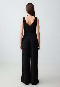Zwarte linnen jumpsuit met een diepe V-rug, brede pijpen, aansluitende taille en een strikceintuur. Glad textuur, minimalistisch ontwerp.