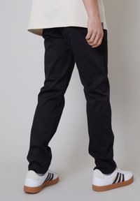 Pantaloni neri in cotone a vestibilità slim, abbinati a sneakers bianche con strisce marroni. La camicia è di colore crema ed è leggermente più lunga sul retro.