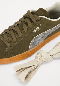 Sněrky olivově zelené z měkkého nubuku s texturovaným stříbrným logem na boku, gumovou podešví a béžovými tkaničkami. Design zahrnuje kulatou špičku a klasický styl.