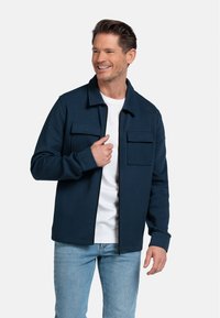 Marineblauwe zip-up jack gemaakt van zachte stof, met een kraag, twee borstzakken en geribbelde manchetten, gedragen met een wit shirt en lichte spijkerbroek.