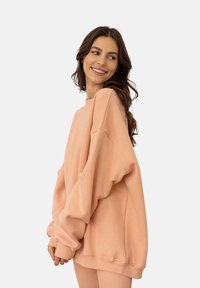 Przytulny, oversizowy sweter w kolorze brzoskwiniowym z ribbowanymi mankietami i dolnym wykończeniem, wykonany z miękkiego materiału. Posiada opadające ramiona oraz luźny krój dla maksymalnego komfortu.