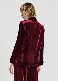 Blazer en velours bordeaux à texture lisse, épaules structurées, et deux boutons sur le poignet, vu de dos.