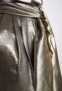 Metalická zlatá sukně s lesklou texturou, širokým pasem se zavázanou mašlí, plisé designem a hladkým, odrazivým povrchem.