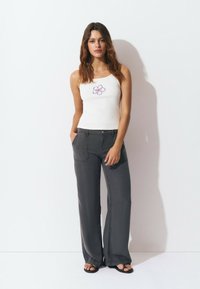 Young woman with long brown hair wearing a white tank top with a purple flower print, gray wide-leg pants, and black sandals, standing.