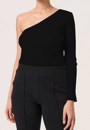 Woman wearing black asymmetrical one-sleeve top and high-waisted black pants, posing with left arm relaxed by side.