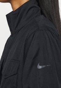 Svart jacka med hög krage, prydd med en silverfärgad Nike-logotyp på ärmen, struttig tyg och en bröstficka.