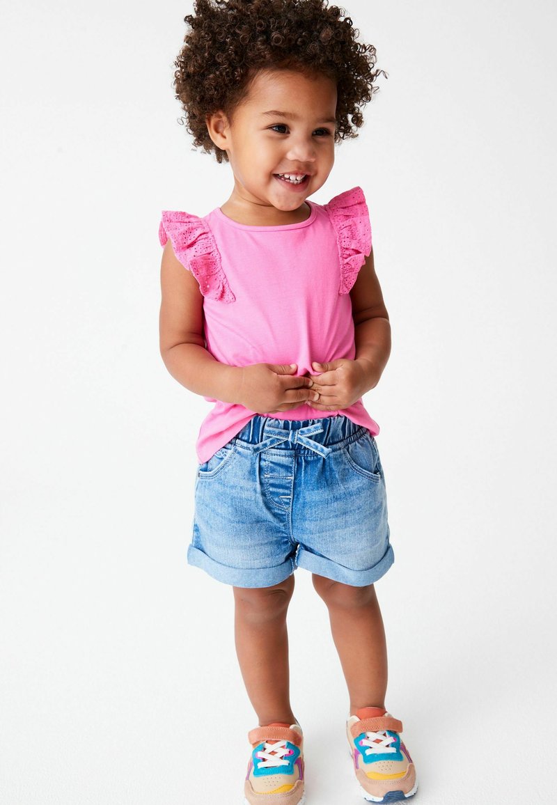 Tout-petit souriant aux cheveux bouclés portant un haut rose à manches volantées, un short en jean et des baskets colorées, debout sur un fond blanc.