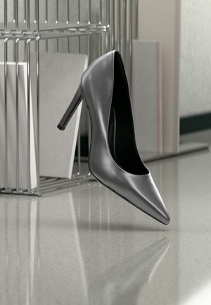 Chaussure noire à talon haut suspendue en l'air devant un classeur métallique avec des dossiers sur une surface polie.