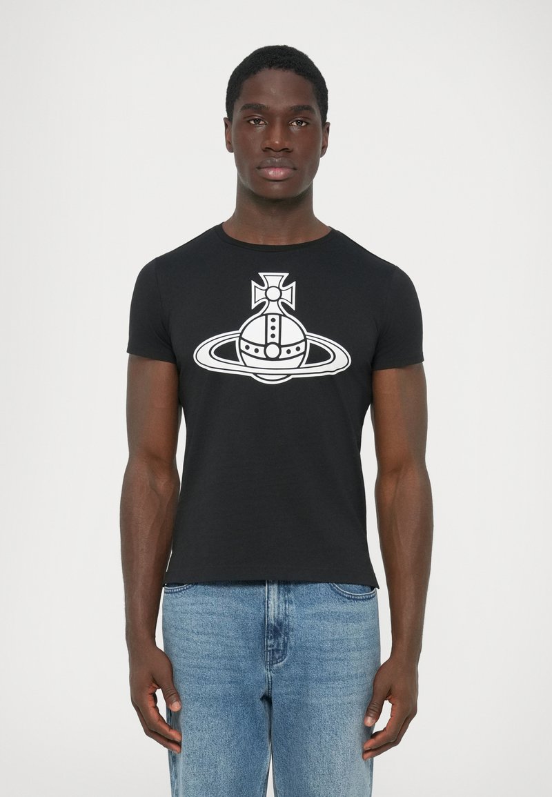 T-shirt nera a maniche corte con grafiche bianche di un pianeta e di una croce, indossata da un giovane in piedi con jeans blu su uno sfondo semplice.