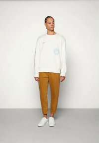 Felpa bianca con grafica blu e logo arancione, abbinata a pantaloni tapered color sabbia e sneakers bianche. Tessuto morbido e vestibilità casual.