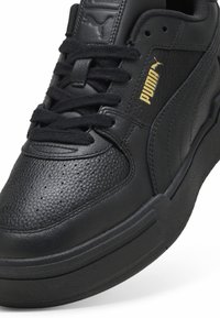 Chaussure de sport noire avec une tige en cuir texturé, des détails perforés, des lacets noirs et un logo de marque doré discret sur le côté.