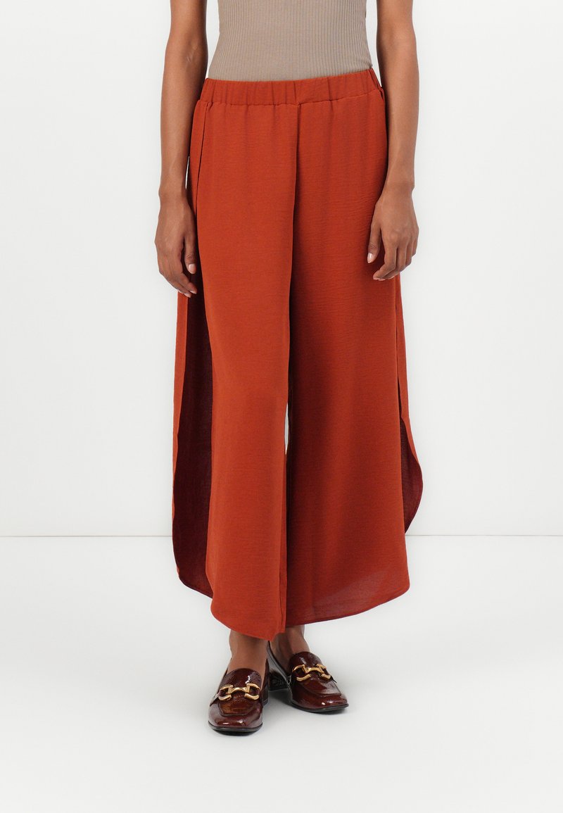Pantalons larges en rust avec un tissu texturé et de hautes fentes sur les côtés, associés à des mocassins marron brillants ornés d'une chaîne en doré.