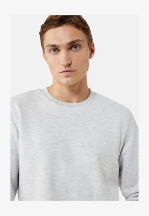 Giovane uomo con capelli castani corti e occhi azzurri che indossa una felpa grigio chiaro con collo rotondo su uno sfondo semplice.