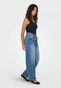 Jeans a gamba larga in denim blu con vita alta, caratterizzati da un'etichetta rossa e bianca sul retro. Indossati con una canotta nera e ballerine nere.