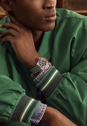 Žmogus dėvi žalią striukę su dryžuotais rankogaliais ir džinsiniais rankovėmis, paguldytą smakrą ant rankos, demonstruojantis sidabrinį rankinį laikrodį su raudonu ir juodu žiedu.