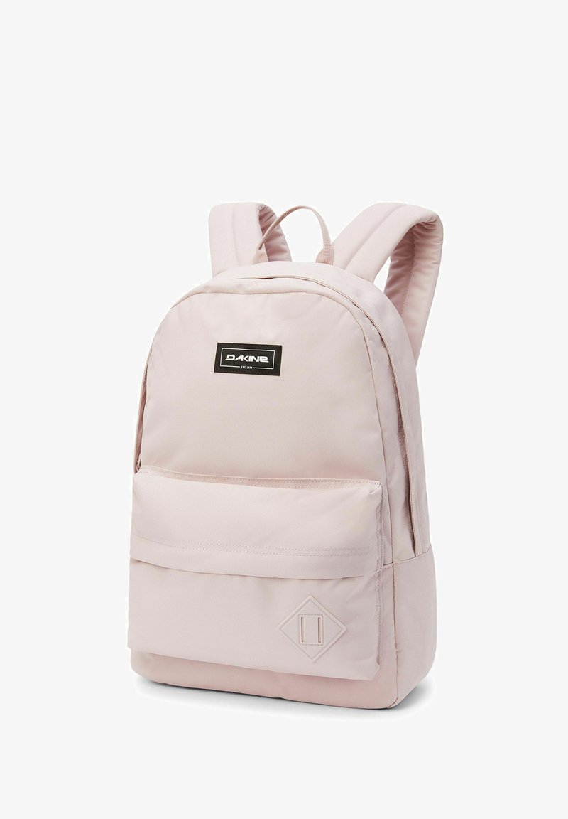 Mochila Dakine de color rosa claro con una textura de tela suave, acento en forma de diamante, bolsillo frontal y correas de hombro acolchadas.