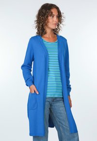 Blauer strukturierter Cardigan mit langen Ärmeln und Taschen, übereinander getragen über einem blau-grün gestreiften Hemd. Jeans runden das Outfit ab.