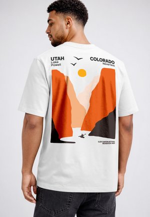 Mann trägt weißes T-Shirt mit orangefarbenem Canyon-Design, Sonne, Vögeln und Text, der auf Utah Lake Powell und Colorado Glen Canyon verweist.