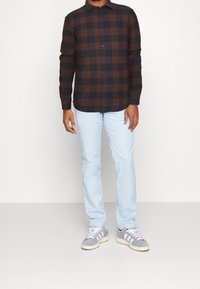 Bruine en zwarte geruite blouse met lange mouwen, knoopsluiting aan de voorkant, gecombineerd met lichtblauwe broek en grijze sportieve schoenen met witte strepen.