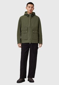 Veste imperméable vert olive avec fermeture éclair à l'avant, capuche détachable, deux poches poitrine et cordons de serrage ajustables. Associée à un pantalon et des chaussures sombres.