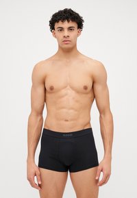 Čierne boxerky SLOGGI s hladkou textúrou, priliehavým dizajnom a elastickým pásom s logom. Model má neutrálnu postúru.