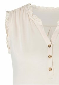 Haut beige clair avec des bords froncés aux manches, un décolleté en V et trois boutons décoratifs en métal doré le long de la patte de boutonnage. Texture de tissu douce.
