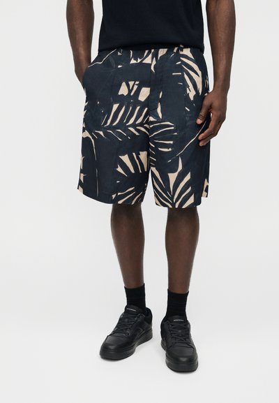 Άνδρας που φοράει μαύρα και μπεζ σορτς με τροπικό σχέδιο, μαύρο μπλουζάκι, μαύρες κάλτσες και μαύρα αθλητικά παπούτσια, στέκεται με τα χέρια στις τσέπες.