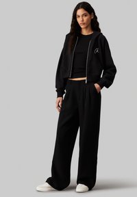 Felpa nera tagliata con zip e tasche frontali, abbinata a pantaloni neri larghi. Le sneakers bianche completano l'outfit, con una texture liscia.
