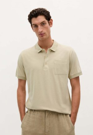 Camisa polo beige de tejido suave, con mangas cortas, cuello y un bolsillo en el pecho. Llevada con pantalones beige claros, contra un fondo neutro.