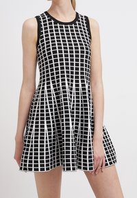 Robe noire sans manches avec motif en grille blanche, ajustée en haut et jupe évasée, portée par une personne sur un fond uni.