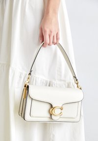 Biała skórzana torebka z klamrą w odcieniu złota, gładką fakturą oraz kontrastującą czarną krawędzią. Do ręki z krótkim paskiem.