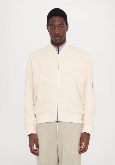 Hellbeige Bomberjacke aus strukturiertem Stoff, mit einem Reißverschluss auf der Vorderseite, seitlichen Taschen und einer Reißverschlusstasche am Ärmel.