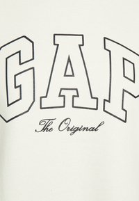 Felpa crema con lettering bold nero "GAP" e un testo più piccolo in corsivo "The Original" sottostante. Texture morbida in cotone.