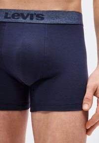 Άνδρας που φοράει σκουρομπλε εσώρουχα Levi’s boxer briefs, με εμφανή τη ζώνη μέσης και το πάνω μέρος του μηρού σε απλό φόντο.