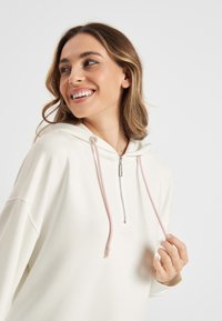 Witte hoodie met roze trekkoorden, ritssluiting bij de kraag en geborduurd logo. Zachte textuur, relaxed fit en brede mouwen. Effen achtergrond.