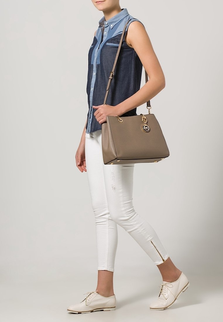 Femme en chemise en denim bleu sans manches, pantalon blanc, chaussures blanches, tenant un sac taupe avec une bandoulière et des accents dorés.