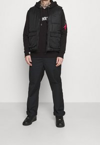 Chaleco acolchado negro con múltiples bolsillos sobre una sudadera negra con capucha, combinado con pantalones negros y zapatos negros, resaltando un estilo en capas.