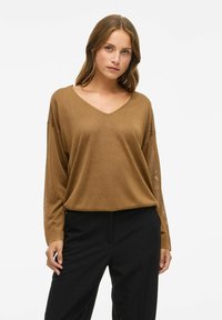 Lichtbruine, lichtgewicht, lange mouw V-hals top met een relaxte pasvorm, voorzien van een zachte textuur en subtiele glans, gecombineerd met zwarte broek.